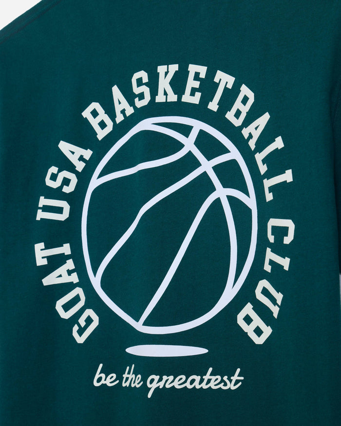 Close-up of a dark teal t-shirt featuring a white graphic print of a basketball and the text 'GOAT USA BASKETBALL CLUB' and 'be the greatest'.