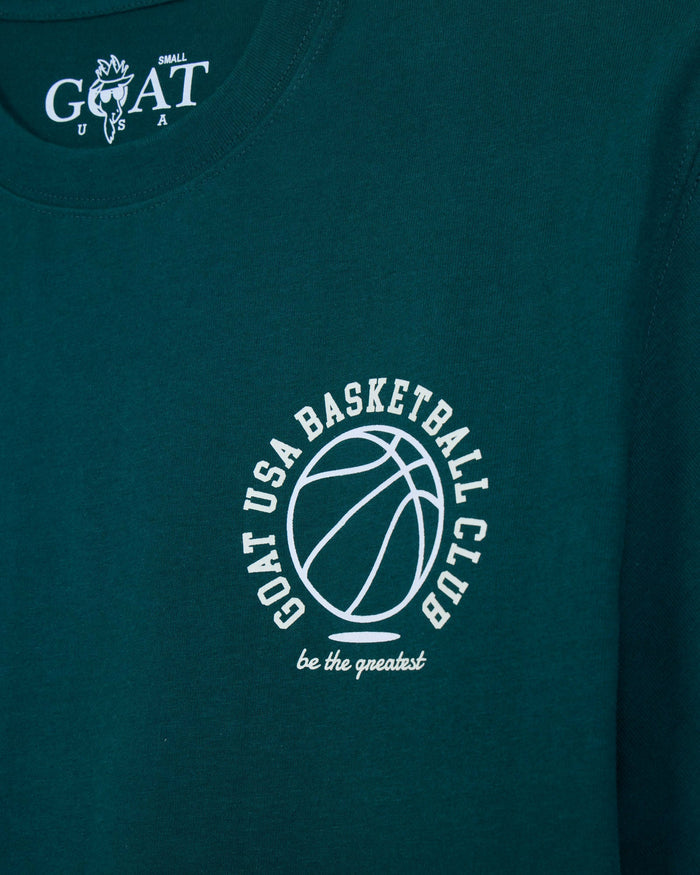 Close-up of a dark teal t-shirt featuring a white circular graphic on the chest with a basketball outline and the text 'GOAT USA BASKETBALL CLUB be the greatest'. The inside collar shows a white 'GOAT' logo.