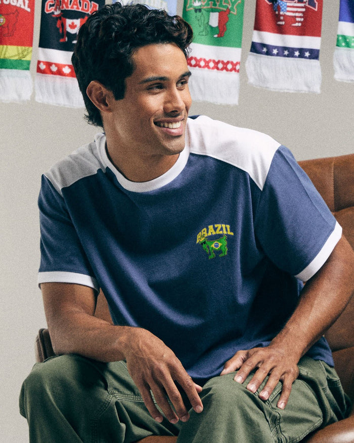 Man wearing a Brazil-themed t-shirt sitting on a couch with flags in the background