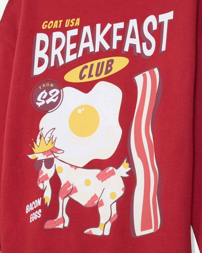 A close-up of a red graphic sweatshirt featuring a cartoon breakfast theme with the text 'GOAT USA BREAKFAST CLUB' and 'BACON EGGS'. The design includes a white goat wearing a yellow crown and sunglasses, a large fried egg, and a striped bacon slice.