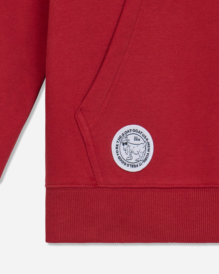 Close-up of a red ribbed sweatshirt sleeve and cuff, featuring a circular white embroidered patch with a goat illustration and text.