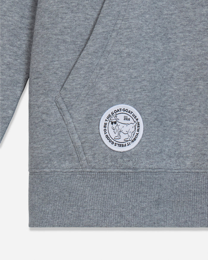 Close-up of a heather gray hoodie showing the left chest area with a circular white and black embroidered patch that reads 'IT FEEELS GOOD TO BE THE GOAT' and features a goat illustration.
