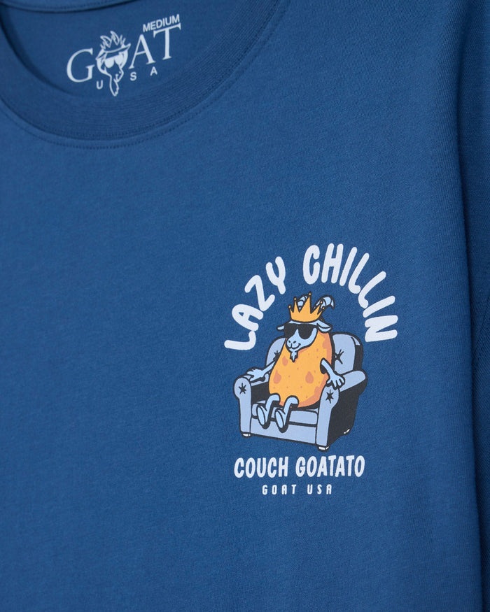 Close-up of a medium blue t-shirt featuring a graphic of a cartoon goat wearing a crown and sitting in a couch, with the text 'LAZY CHILLIN' and 'COUCH GOATATO GOAT USA' printed around it.