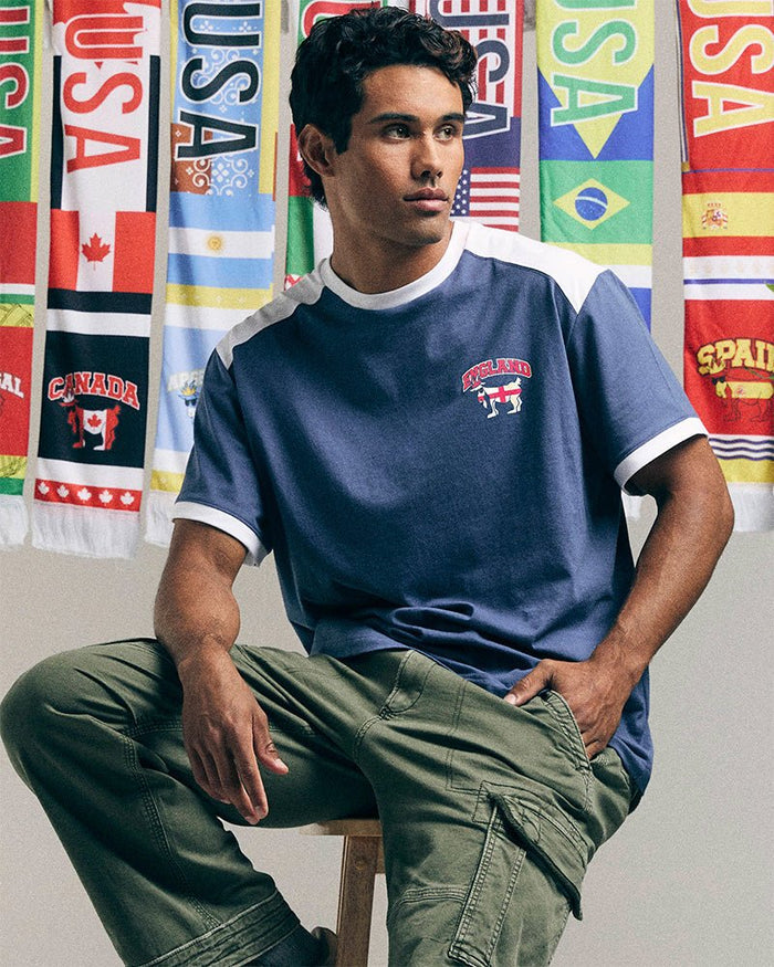 Man sitting on a stool wearing a blue t-shirt with a logo and green pants, with colorful flags in the background.
