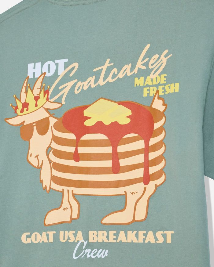A close-up of a sage green t-shirt featuring a graphic of a goat wearing sunglasses and a crown, with a stack of pancakes topped with syrup and butter. The text reads 'HOT Goatcakes MADE FRESH' and 'GOAT USA BREAKFAST Crew'.