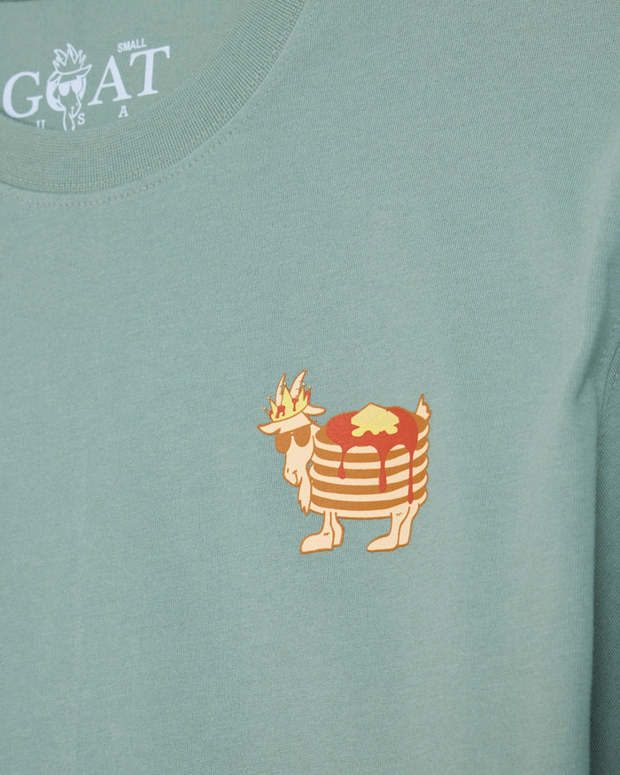 A close-up of a sage green t-shirt featuring a small graphic of a character resembling a flaming pancake on the chest, with the white 'GOAT' brand logo printed inside the collar.