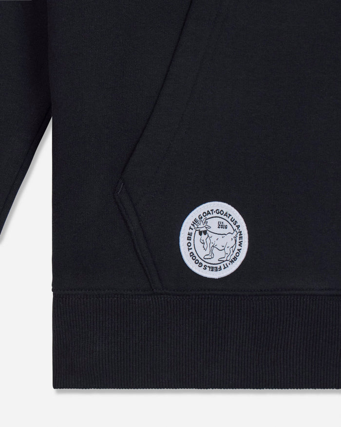 Close-up of a black ribbed sweatshirt cuff with a circular white embroidered logo patch featuring a bear and the text 'IT FEELS GOOD TO BE THE GOAT'.