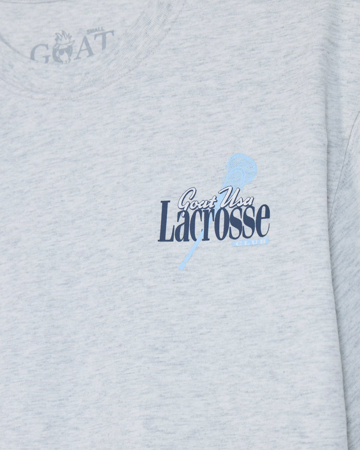 Close-up of a light heather gray t-shirt featuring a navy blue and light blue 'Goat USA Lacrosse Club' graphic on the chest, with a faint 'GOAT' logo printed inside the collar.