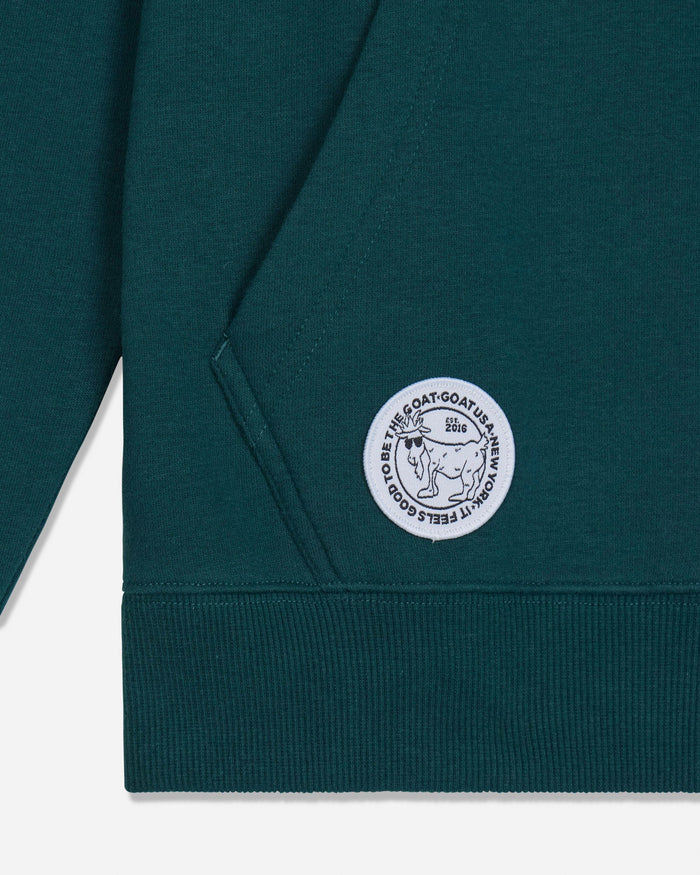 Close-up of a dark teal-green sweatshirt showing the sleeve, ribbed cuff, and a circular white embroidered patch on the pocket with the text 'IT FEELS GOOD TO BE THE GOAT' and a goat illustration.
