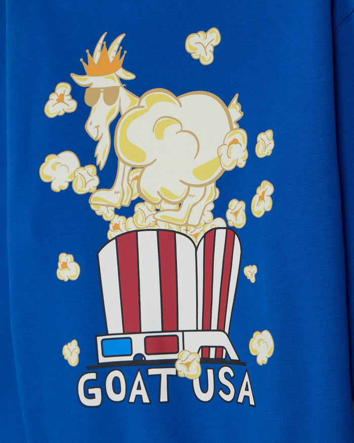 A close-up of a royal blue t-shirt featuring a graphic print of a cartoon goat wearing sunglasses and a crown, popping popcorn out of a red and white striped bucket, with the text 'GOAT USA' below.
