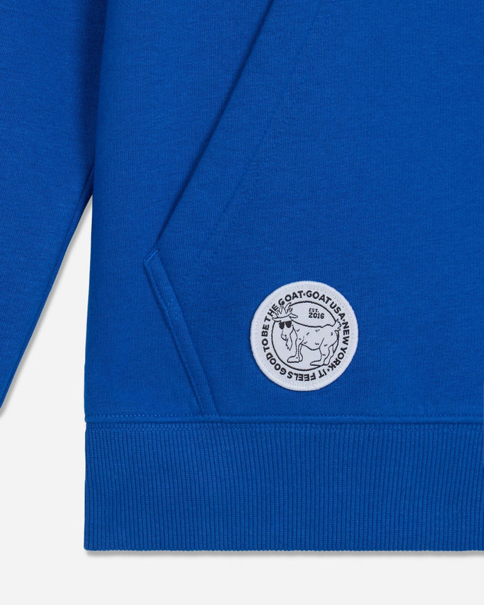 Close-up of a royal blue sweatshirt showing the ribbed cuff, a diagonal seam, and a circular white embroidered patch featuring a goat illustration and the text 'IT FEELS GOOD TO BE THE GOAT'.