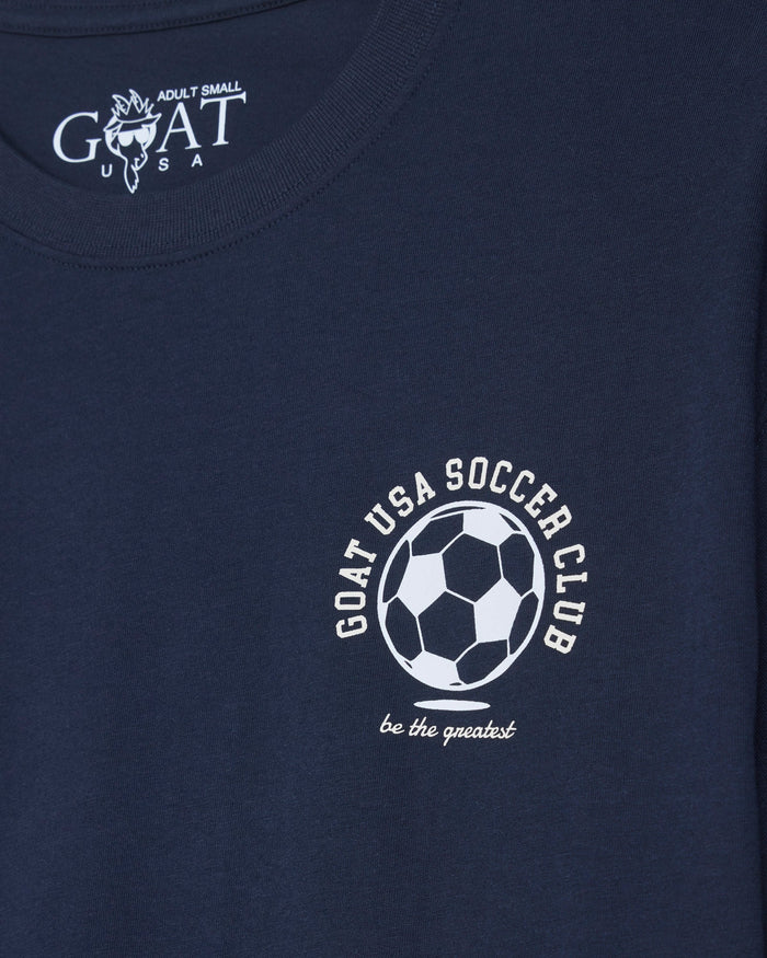 Close-up of a navy blue t-shirt featuring a white printed graphic of a soccer ball with the text 'GOAT USA SOCCER CLUB' and 'be the greatest' below it.