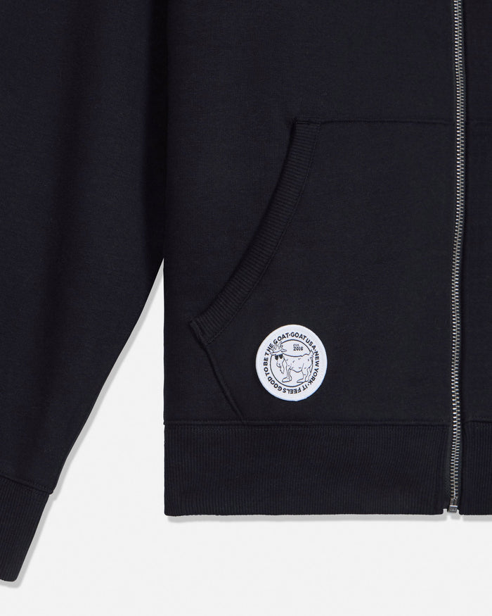 Close-up of a black zip-up hoodie showing the left chest pocket, ribbed hem and cuff, and a circular white embroidered patch with a goat and text.#color_black