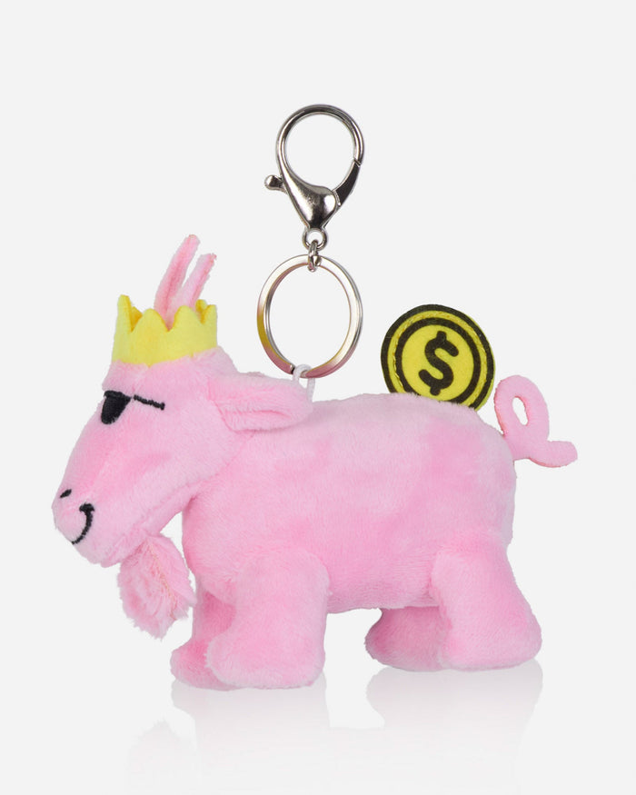 A pink plush goat keychain charm with a yellow crown, black embroidered eyes and mouth, a silver metal clasp, and a yellow coin-shaped tag with a dollar sign.