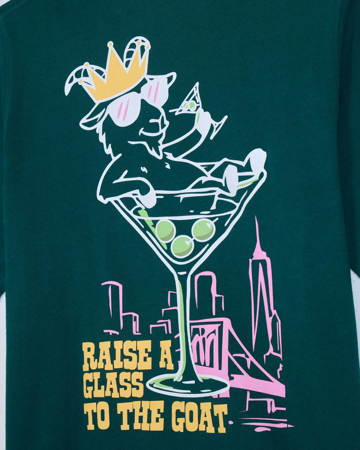 Close-up of sage t-shirt with martini glass goat graphic and text that reads 'Raise a glass to the GOAT'