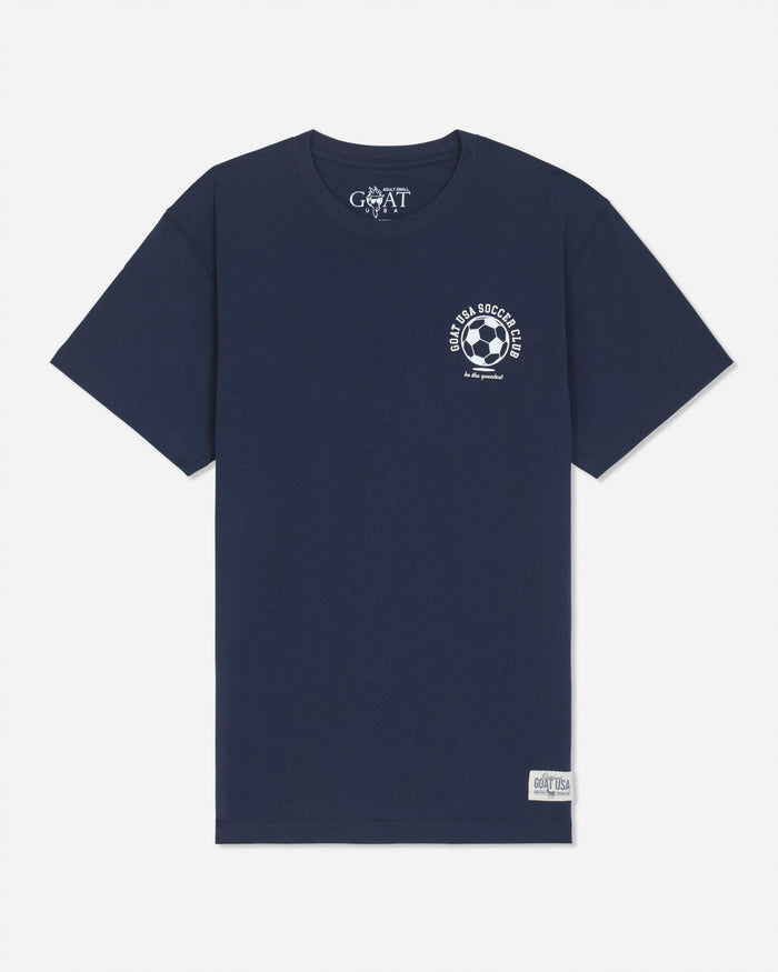 A navy blue short-sleeve t-shirt with a white circular soccer-themed graphic on the left chest and a white 'GOAT' brand print inside the collar, displayed on a light gray background.
