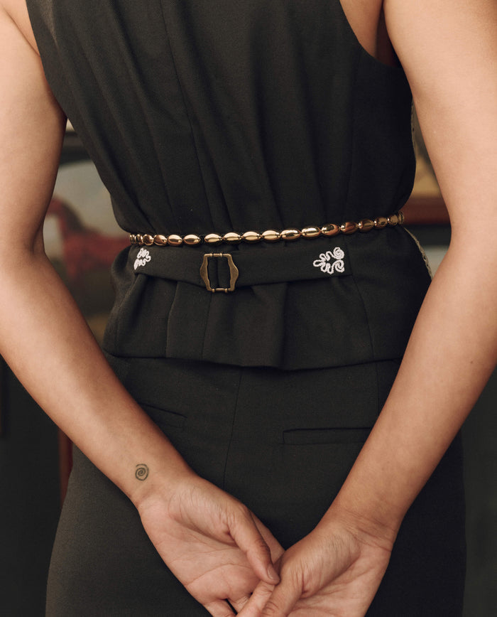 A person wearing a sleeveless black dress with THE GREAT. F26 STETSON’s The Beaded Belt in Black With Gold—highlighting Western craftsmanship—stands with hands clasped behind their back; a small spiral tattoo is visible on their left wrist.