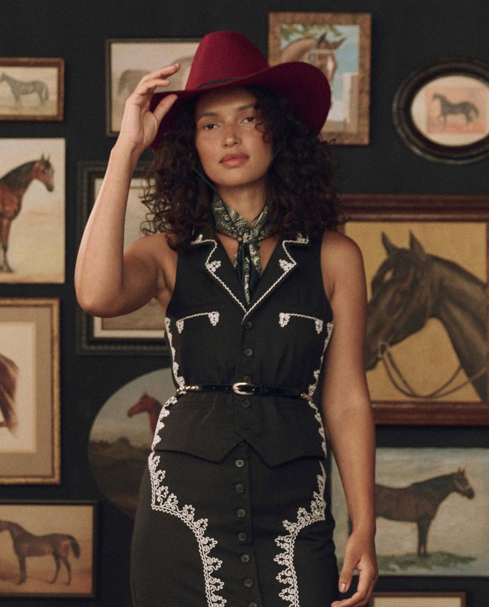 A woman in a black Western-style dress and red cowboy hat stands before a wall adorned with framed horse paintings, her outfit highlighted by THE GREAT. F26 STETSON The Beaded Belt -- Black With Gold, a piece that exemplifies exceptional Western craftsmanship.