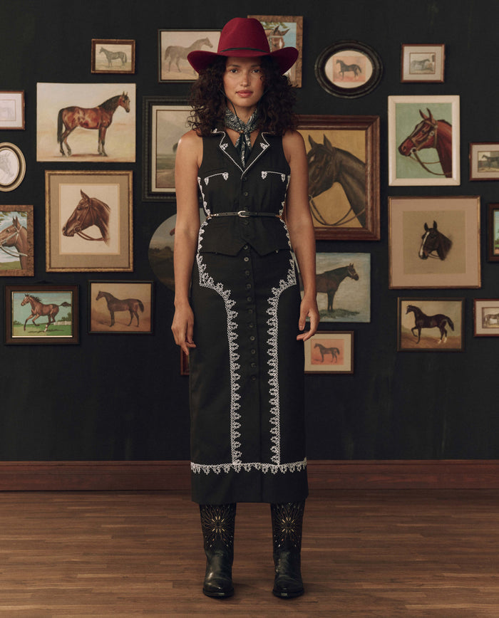 Woman in a black Western-style dress, red hat, and cowboy boots stands before a wall adorned with framed horse art, her look completed by THE GREAT. F26 STETSON The Beaded Belt in Black With Gold—a piece that highlights classic Western craftsmanship.