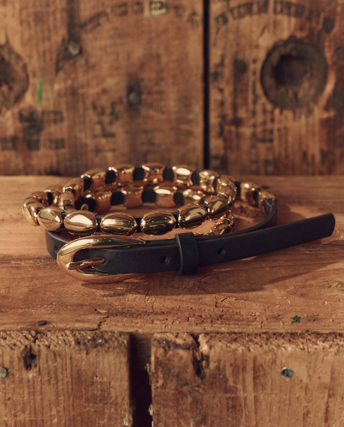 The Beaded Belt — Black With Gold by THE GREAT. F26 STETSON is elegantly coiled on a rugged wooden surface with a rustic wooden backdrop, highlighting its top-grain black leather, striking gold oval links and buckle, and exceptional Western craftsmanship.