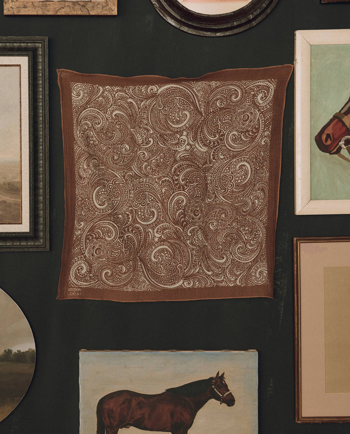 The Stetson Western Paisley Scarf in rust by THE GREAT. F26 STETSON—a brown square scarf featuring a white paisley pattern inspired by vintage bandanas—is displayed on a dark wall, surrounded by framed artwork and paintings, including images of horses.