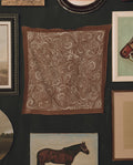 The Stetson Western Paisley Scarf in rust by THE GREAT. F26 STETSON—a brown square scarf featuring a white paisley pattern inspired by vintage bandanas—is displayed on a dark wall, surrounded by framed artwork and paintings, including images of horses.