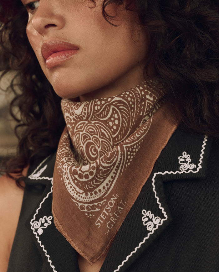 A person wearing The Stetson Western Paisley Scarf in Rust by THE GREAT. F26 STETSON, paired with a black vest featuring white embroidered details.