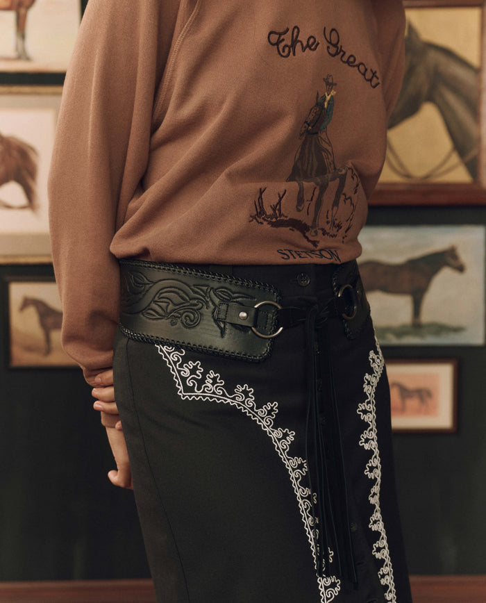 A person wearing a brown sweater featuring a cowboy graphic and an embroidered black skirt, accessorized with THE GREAT. F26 STETSON The Tooled Corset Belt in Black, stands in front of framed horse paintings.