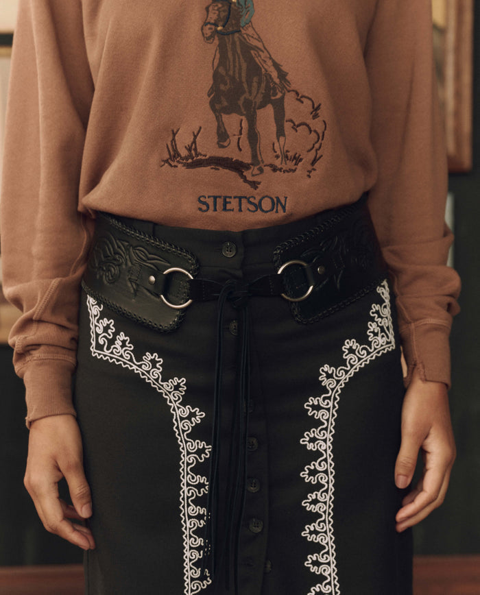 Wearing a brown THE GREAT. F26 STETSON shirt with a horse graphic, paired with a black skirt featuring white embroidery, and styled with The Tooled Corset Belt in black—complete with classic western accessories.