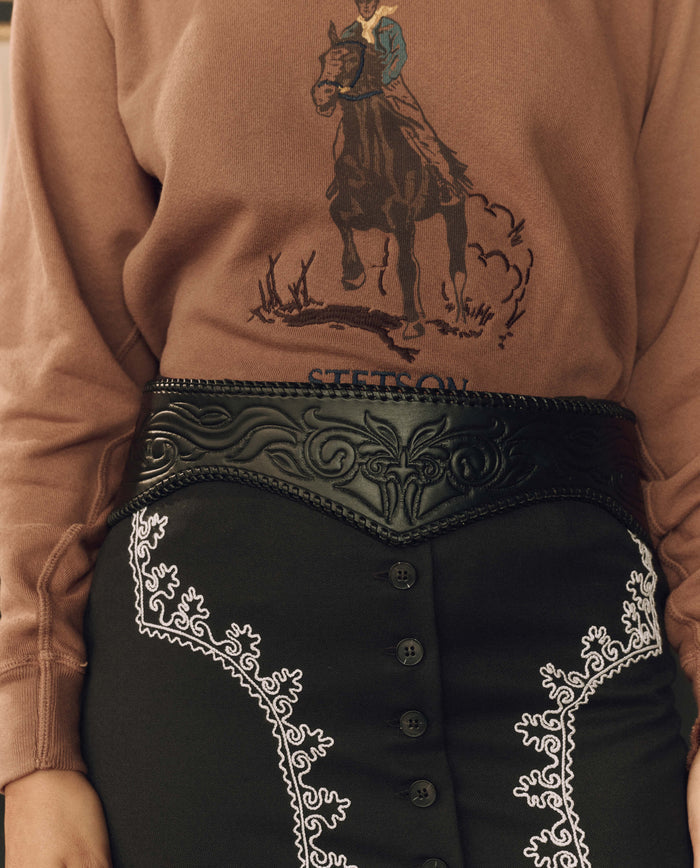 Wearing a brown sweatshirt with a horse rider graphic and a black skirt adorned with white embroidery, this look is finished with THE GREAT. F26 STETSON’s The Tooled Corset Belt in black for an added touch of western flair.