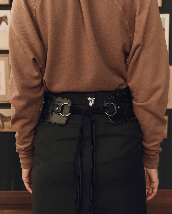 A person facing away wears a brown sweater and black skirt, accessorized with THE GREAT. F26 STETSON The Tooled Corset Belt in black. Framed pictures hang on the wall in the background.