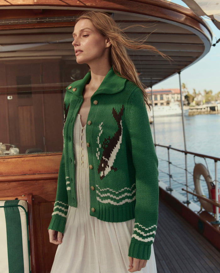 A woman wearing THE GREAT. SU26's The Bass Cardigan in Grove, featuring a whale pattern, stands on a wooden boat with water and buildings in the background.