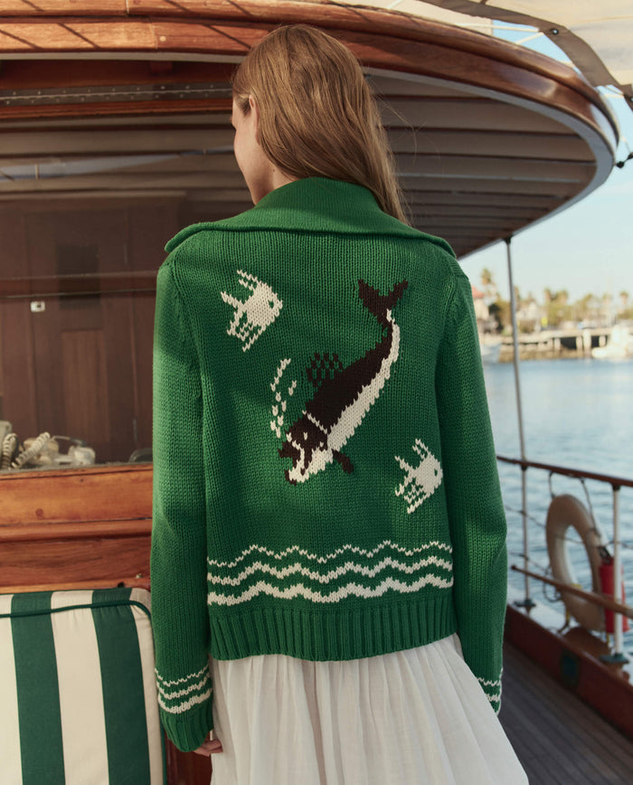 A person wearing THE GREAT. SU26 The Bass Cardigan -- Grove, featuring a whale and fish design on the back, stands on a boat facing away from the camera.