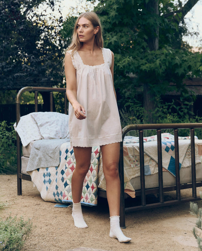 A woman wears The Great. SP26 Sleep Eyelet Mini Tank Dress in True White and stands outdoors by a metal-framed bed with colorful quilts, surrounded by trees and greenery.