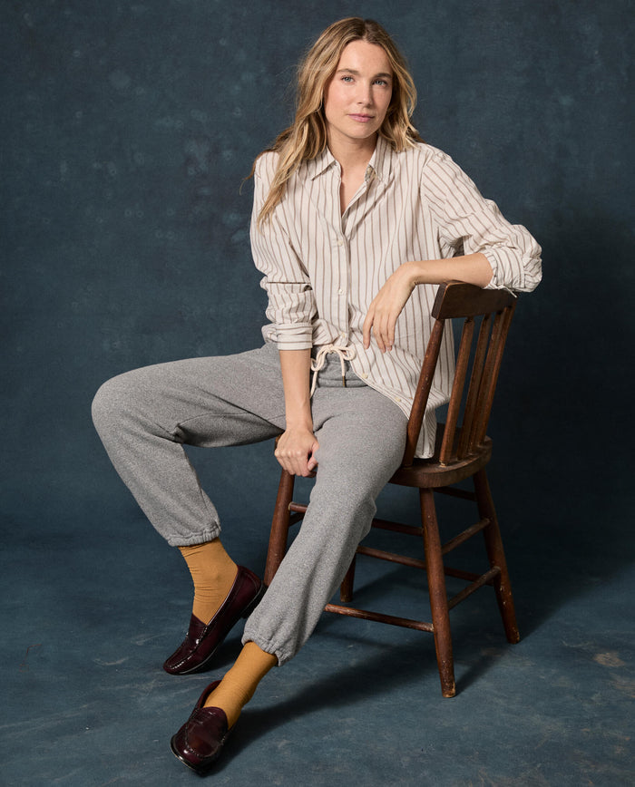 A person sits on a wooden chair against a dark backdrop, wearing a striped shirt, yellow socks, burgundy loafers, and The Fleece Stadium Sweatpant in Dark Heather Grey by THE GREAT. F26 D1 FLEECE BACK KNITS.