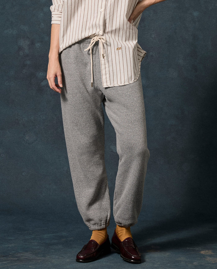 Wearing THE GREAT.’s Fleece Stadium Sweatpant in Dark Heather Grey, a person in a striped button-up shirt, yellow socks, and brown loafers stands against a dark background.