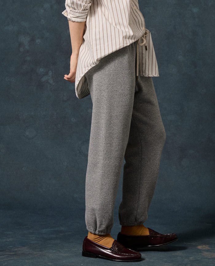 Certainly! Here’s the revised sentence using your product data: Person standing sideways wearing a beige striped shirt, yellow socks, brown loafers, and The Fleece Stadium Sweatpant in Dark Heather Grey by THE GREAT. F26 D1 FLEECE BACK KNITS against a dark textured background.
