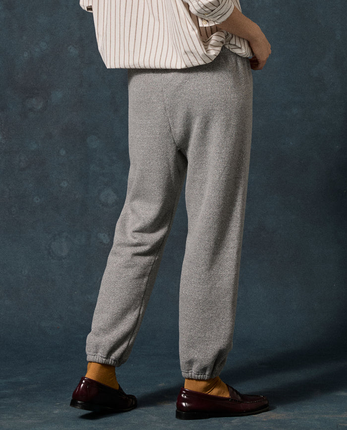 A person stands facing away, wearing a striped shirt, THE GREAT. F26 D1 FLEECE BACK KNITS The Fleece Stadium Sweatpant in Dark Heather Grey, mustard yellow socks, and dark brown loafers, against a dark blue textured background.