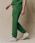 Person wearing THE GREAT. SU26 SEASONAL CORE The Jersey Jogger Pant in Fennel, with a matching green top and white high-top Converse sneakers, standing against a neutral fabric backdrop.