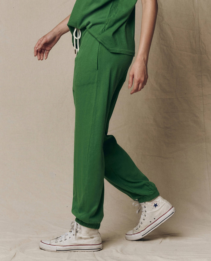Person wearing THE GREAT. SU26 SEASONAL CORE The Jersey Jogger Pant in Fennel, with a matching green top and white high-top Converse sneakers, standing against a neutral fabric backdrop.