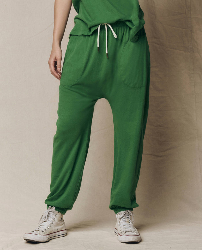 A person wears The Jersey Jogger Pant in Fennel by THE GREAT. SU26 SEASONAL CORE with white sneakers, standing against a plain beige background.
