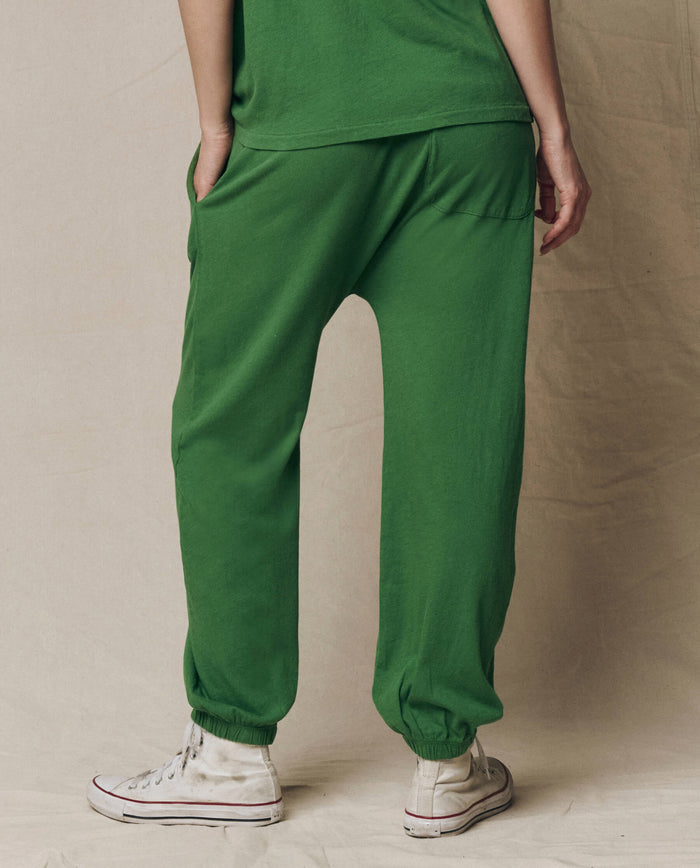Model wearing THE GREAT. SU26 SEASONAL CORE The Jersey Jogger Pant in Fennel with a matching green top and white high-top sneakers, standing with hand in pocket, facing away from the camera against a plain background.