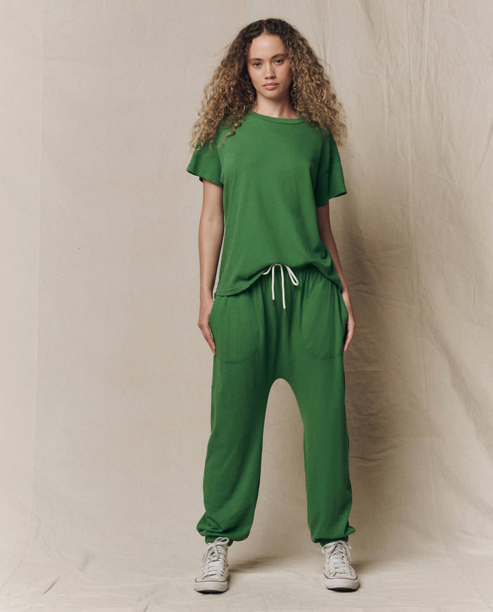 A person faces forward wearing THE GREAT. SU26 SEASONAL CORE The Jersey Jogger Pant in Fennel with a matching tee and white sneakers, set against a plain beige backdrop.
