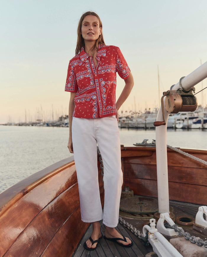 A woman stands on a wooden boat, wearing THE GREAT. SU26’s The Postcard Shirt and the Red Sailboat Bandana. White pants and black sandals complete her look, while a marina with docked boats appears in the background.