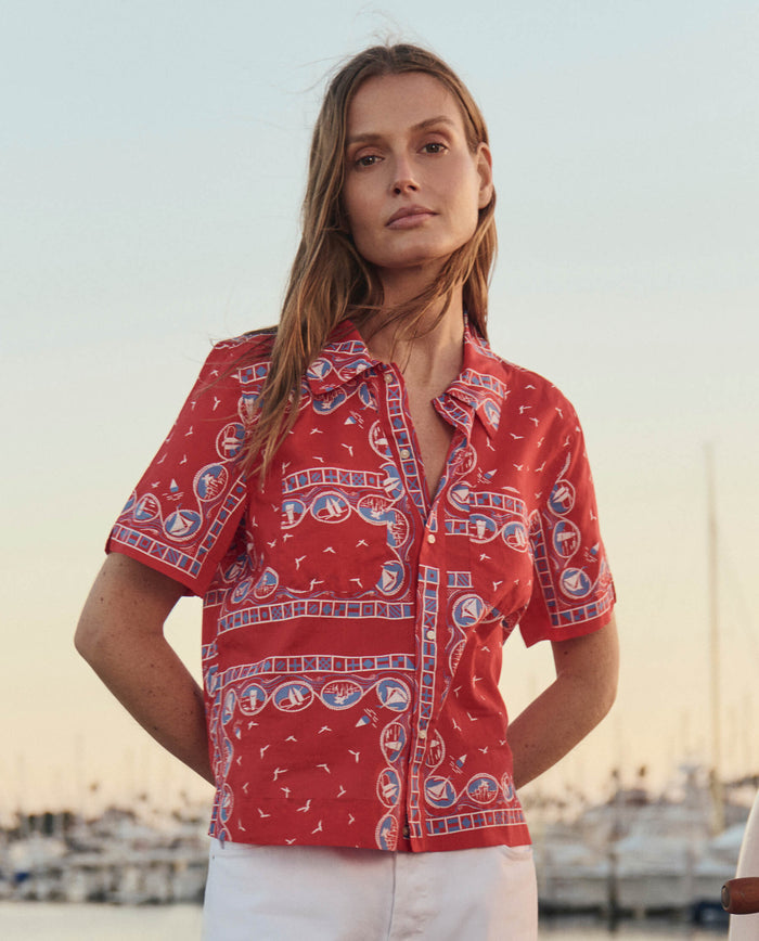 A person stands outdoors by a marina in THE GREAT. SU26’s The Postcard Shirt – Red Sailboat Bandana, paired with white pants, with sailboats in the background.