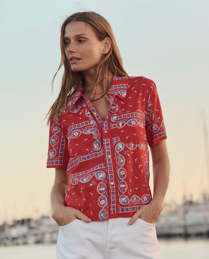 A woman stands outdoors near a marina at sunset wearing THE GREAT. SU26’s The Postcard Shirt in Red Sailboat Bandana and white pants.