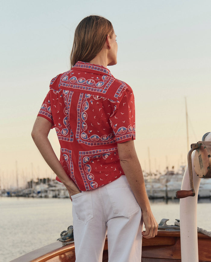 A person in THE GREAT.'s The Postcard Shirt, styled with a Red Sailboat Bandana, stands on a boat facing away, with white pants and a marina of sailboats in the background.