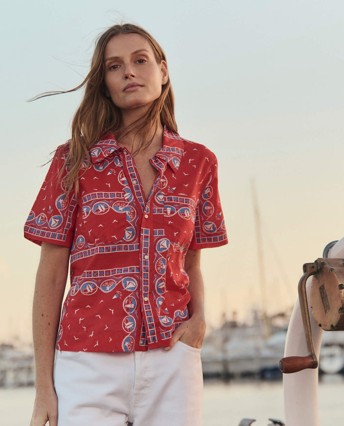 A woman stands outdoors near a marina, wearing THE GREAT. SU26’s The Postcard Shirt in Red Sailboat Bandana with white pants; boats and water are in the background.