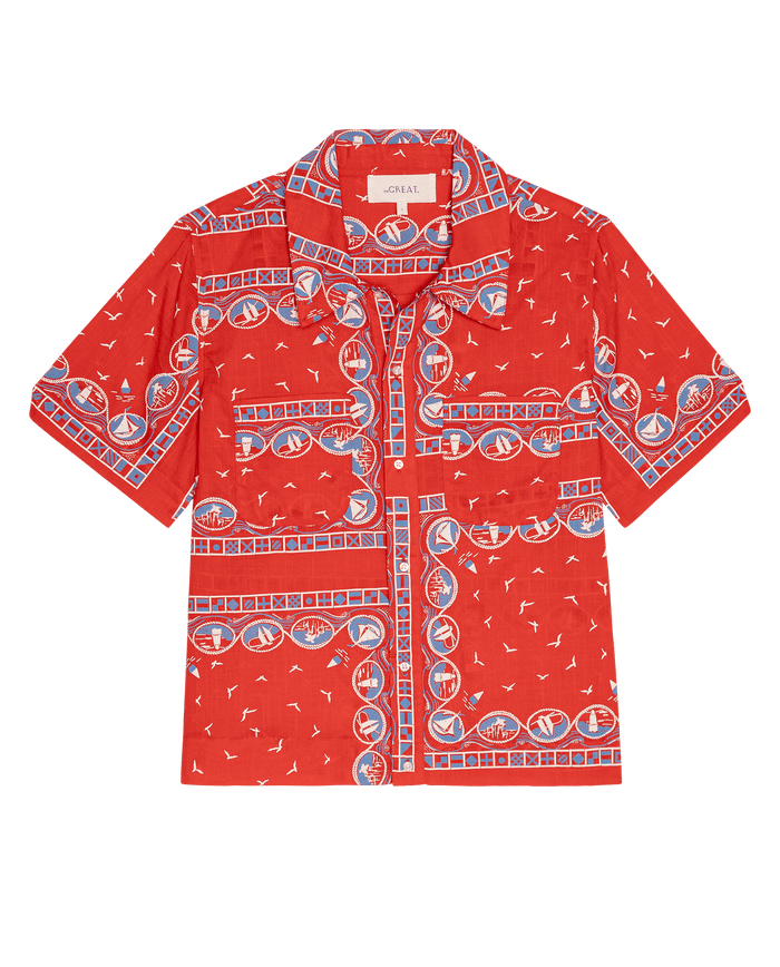 THE GREAT. SU26 The Postcard Shirt – Red Sailboat Bandana: a short-sleeve, button-up shirt with a red background and blue, white, and tan geometric and nautical designs.