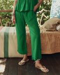 Wearing THE GREAT. SP26 SWIM's The Reef Pant in Bright Sprig, a person stands by a bed with a tan quilt and striped blanket on a wooden deck, surrounded by greenery.
